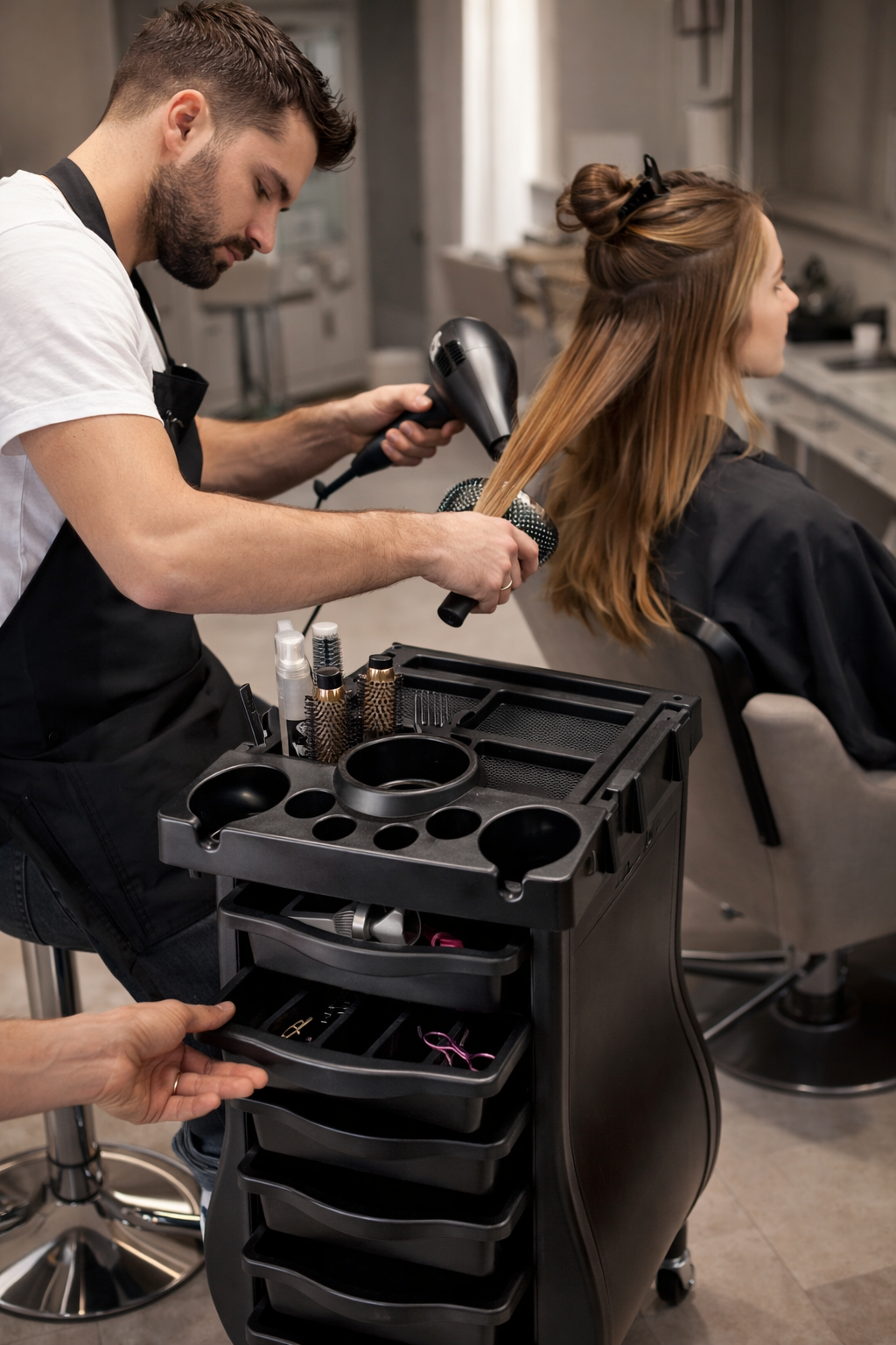 Hairdresser using a hair dryer on a client with a black hair styling cart in a salon.